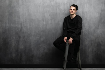 Studio fashion portrait of young sexy man in black hoodie and jeans. Guy sits on a high bar stool against a gray textured wall with free copy space