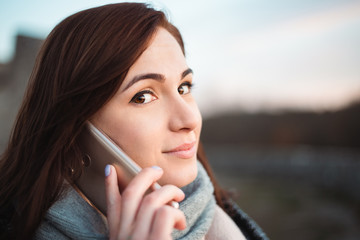 Portrait of young beautiful smilling woman with big eyes calling with cell telephone -  captured from side (profile). Brunette model (girl) looking to camera and calling with phone in nature.