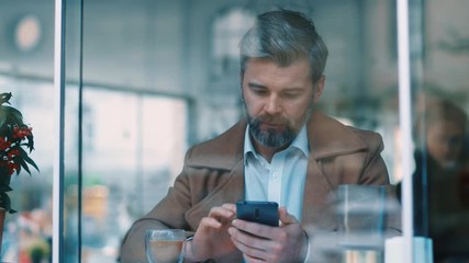 Wall Mural - View through the window of attractive serious middle aged businessman using phone at coffee shop texing message working watching video successful people daily routine entertaining communicating