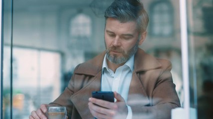 Wall Mural - View through the window of handsome serious middle aged businessman uses phone drinks coffee at coffee shop texing message working successful people daily routine entertaining relaxing communicating