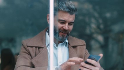 Wall Mural - Close up rotation view of gorgeous middle aged beared business man with beige coat uses phone drinks coffee at cafe texing message working successful people relaxing new technologies communicating