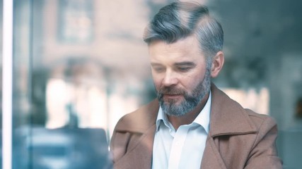 Wall Mural - Close up view through the window of handsome middle aged businessman works drinks coffee smile in cafe. Man use laptop. Business and entrepreneurship successful confident social network apps portrait