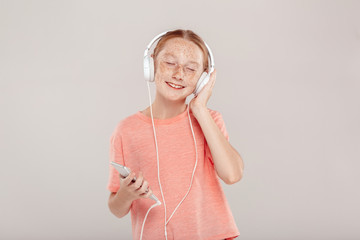 Wall Mural - Inclusive Beauty. Girl with freckles in headphones standing isolated on grey holding smartphone listening music closed eyes delightful