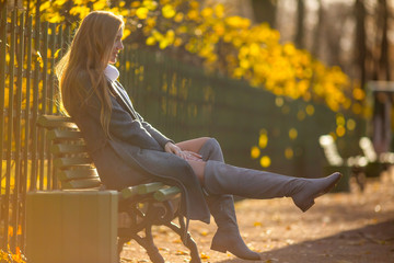 Beautiful girl with light brown hair is sitting on a bench in the alley of an autumn park. Attractive young woman in a gray coat and high boots. Fall season. Golden leaves. Amazing autumn mood.