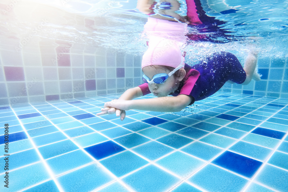 Underwater young little cute girl is swimming in the swimming pool with her swimming teacher. Seen under water while she is diving forward ahead of her. Swimming kid in the pool concept. - obrazy, fototapety, plakaty 