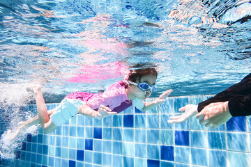 Underwater young little cute girl is swimming in the swimming pool with her swimming teacher. Seen under water while she is diving forward ahead of her with coach. Swimming kid in the pool concept.