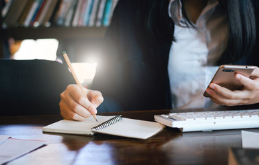 Close-up of hand young businesswoman using mobile phone working with computer and writing information on notebook