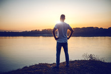 Rear View Of Handsome Caucasian Man Standing With Hands On Hips On Cliff And Looking At River And Beautiful Sunset.