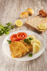 Preparation of a healthy battered fish with egg and breadcrumbs cooked in the oven. It is seasoned with fresh parsley and lemon and accompanied by cooked potatoes and tomatoes.