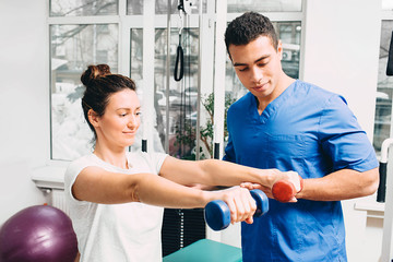 woman correctly performs exercise in the gym for the treatment of arthritis of the hands. The physiotherapist oversees the patient’s exercise. Two dumbbells in the hands of a woman for exercise