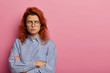Photo of offended displeased angry woman stands with insulted look and crossed hands, sulks while looking away, wears round glasses and blue striped shirt, isolated over rosy wall, copy space