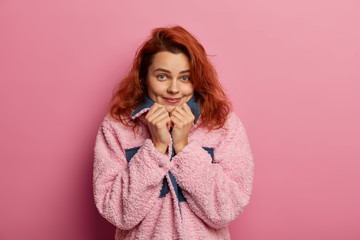 Wall Mural - Pretty pleased ginger haired girl feels comfort wearing new pink warm coat, looks directly at camera, has dimples on cheeks, poses against rosy background, feels shy in presence of handsome man