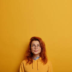 Wall Mural - Vertical image of lovely ginger girl looks above, has pleased expression, raises head, enjoys sunny day, wears big glasses and sweatshirt, isolated over yellow background, copy space area for advert