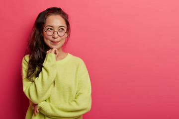 Wall Mural - Shot of attractive thoughtful girl recalls first romantic date with boyfriend, keeps hands gently under chin, has dreamy happy expression, wears casual clothes, models over rosy wall, copy space