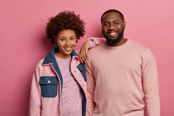 Photo of friendly happy woman and man with dark skin stand closely, smiles happily, wear fashionable pastel clothes, look at camera positively, isolated on pink background. Afro American couple