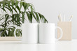 Two mugs mockup with workspace accessories on a white table and a palm plant.