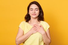 Image Of Satisfied Generous Brunette Woman With Charming Smile, Keeps Both Palms On Chest, Wearing Casual Yellow T Shirt, Standing Against Studio Wall, Being Kind Hearted. People, Gratitude Concept.