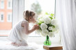 © alexkich - A little girl is sitting on the windowsill. A bouquet of flowers in a vase by the window and a girl sniffing flowers. A little princess in a white dress with a bouquet of white flowers by the window.