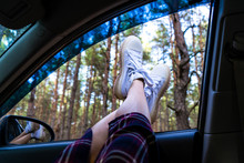 Enjoyment, Relaxation In The Forest, Lazy Sunday, Tourism, Travel, Free Time, Rest, The Concept Of Happiness. Legs Of A Young Girl In White Boots In A Car Window On A Background Of Coniferous Forest.