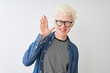 © Krakenimages.com - Young albino blond man wearing denim shirt and glasses over isolated white background Waiving saying hello happy and smiling, friendly welcome gesture