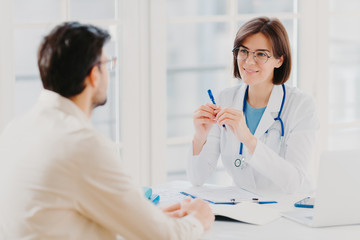 Male patient visits doctor at clinic, tells about his problems, talk about illness. Attentive female doctor writes down notes in medical history, makes prescriptions, listens to patients health issues