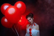 Portrait Cute Young Woman Crazy Smiling Face Clown In White Costume Stands Backdrop Dark Gothic Room Holding Red Balloons Evil Eyes. Art Creative Bright Halloween Make-up Hairstyle. Fog Smoke Party 