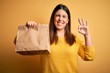 Young beautiful woman holding take away paper bag from delivery over yellow background doing ok sign with fingers, excellent symbol