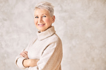 People and aging concept. Portrait of beautiful elegant middle aged woman in warm cozy turtle neck sweater smiling, keeping arms crossed on chest, her posture and look expressing confidence