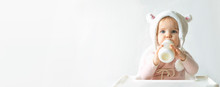 Little Toddler Girl In A Warm Fluffy Hat Drinks Milk From A Bottle While Sitting. Half-length Portrait. White Gray Background. Banner, Free Space, Copy Space.