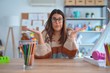 Young beautiful teacher woman wearing sweater and glasses sitting on desk at kindergarten clueless and confused expression with arms and hands raised. Doubt concept.