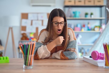 Sticker - Young beautiful teacher woman wearing sweater and glasses sitting on desk at kindergarten feeling unwell and coughing as symptom for cold or bronchitis. Healthcare concept.