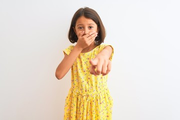 Young beautiful child girl wearing yellow floral dress standing over isolated white background laughing at you, pointing finger to the camera with hand over mouth, shame expression