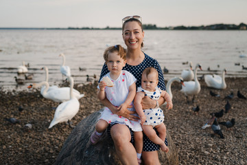 Young mother with her baby girl daughters feeding swan and little ducklings birds bread at a river wearing dotted dress - Family values warm color summer scene