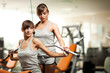 © YouraPechkin - Young beautiful brunette woman in sportswear helping another girl with using fitness machine for back in gym. Healthy active lifestyle, sport, bodycare concept