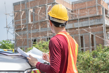 Thai young male engineer or architect wearing yellow-orange vest and yellow safety helmet is looking at blueprint paper placing in a car in front of unfinished house.