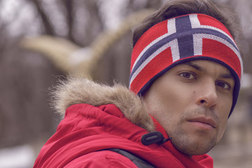 Handsome man athlete in a red jacket and a hat on his head with the flag of Norway, in winter, turned his head back looking into the frame