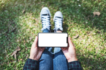 Top view mockup image of a woman holding black mobile phone with blank white screen while sitting in the outdoors