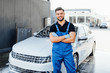 Professional bearded washer in blue uniform washing luxury car stands near car in bubbles.