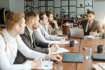 busy caucasian men in tux sit on table in office listening to boss and make notes in notebook, attentively listen to advices and strategies for future work by leader