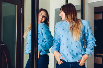 Wall Mural - Woman in sweater looking at mirror. Girl trying clothes on by wardrobe at home. Beauty fashion