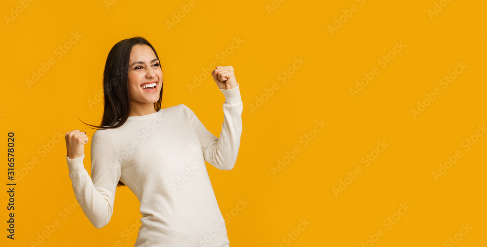 Euphoric brunette woman celebrating success with raised fists over yellow background - obrazy, fototapety, plakaty 