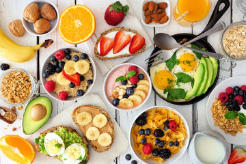 Wall Mural - Healthy breakfast table scene with fruit, yogurts, oatmeal, smoothie bowl, cereal, nutritious toasts and egg skillet. Top view over a white wood background.