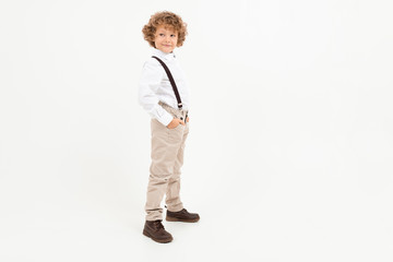 Charming boy with curly hair in white shirt with black suspenders stands isolated on white background