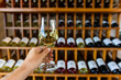© Valmedia - hand holding a glass of white wine selective focus view, tasting room wines bottles display on wooden racks shelves background, wine shop interior
