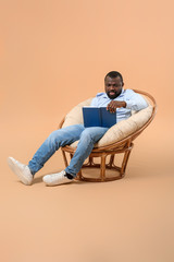 Poster - African-American man reading book while sitting in armchair against color background