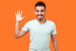 Hello! Portrait of friendly glad handsome brunette man with beard in casual white t-shirt showing hi gesture with waving hand and smiling sincerely. indoor studio shot isolated on orange background