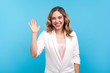 Hello! Portrait of friendly kind cheerful girl with wavy hair in white jacket waving hand saying hi welcome, smiling with hospitable sociable expression. indoor studio shot isolated on blue background