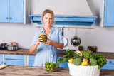 Fototapeta  - Young pretty woman holding jar of pickles and smiling looking at camera, cooking green salad in modern kitchen, preparing vegetarian meal, basket of fresh vegetables on table, healthy food, vitamins