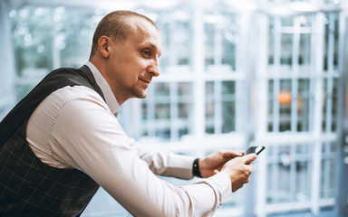 Wall Mural - An elegant mature cheerful businessman using his cellphone while leaning against a chrome railing of a balcony indoors of a modern office open-space area, with a copy space place on the right