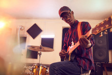 Low Angle Portrait Of Contemporary African-American Man Playing Guitar And Looking At Camera During Rehearsal Or Concert With Music Band In Studio, Copy Space